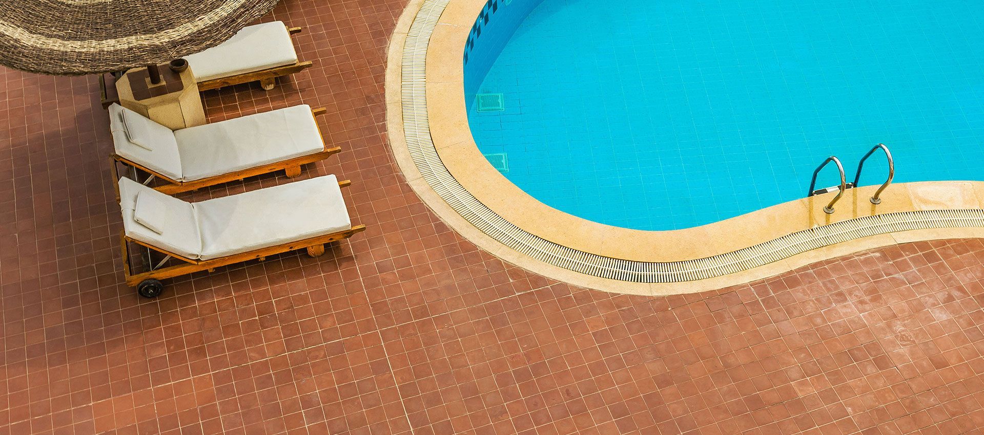 Top-down view of three white lounge chairs beside a turquoise swimming pool on a brown tiled patio.