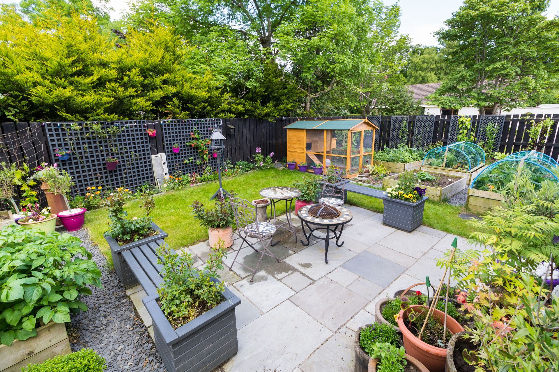 A small garden shovel rests against a wooden crate filled with colorful flowers in a garden bed with mulch and rocks.
