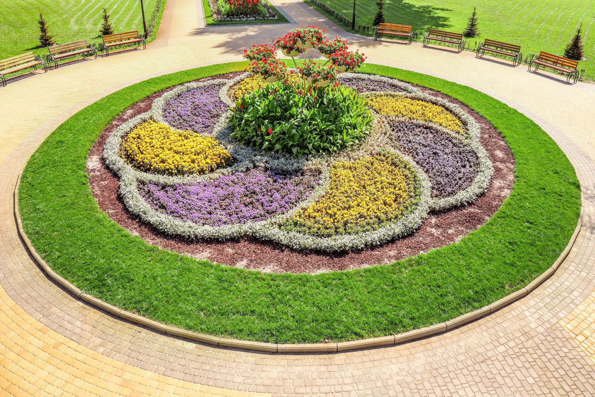 A top-down view of a circular garden bed with swirling, colorful flower patterns, surrounded by a lawn and benches.