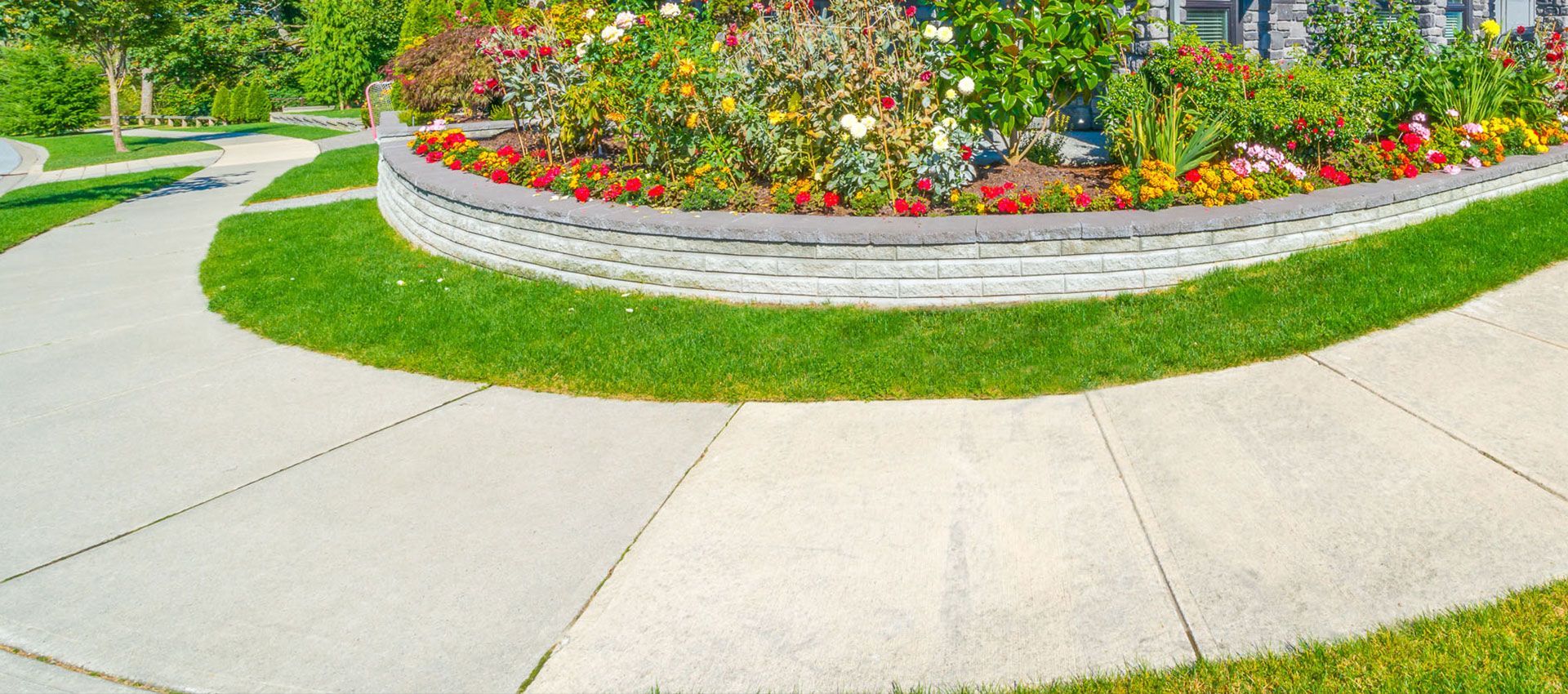 A curved concrete sidewalk borders a raised flower bed filled with colorful blooming plants and green shrubs.