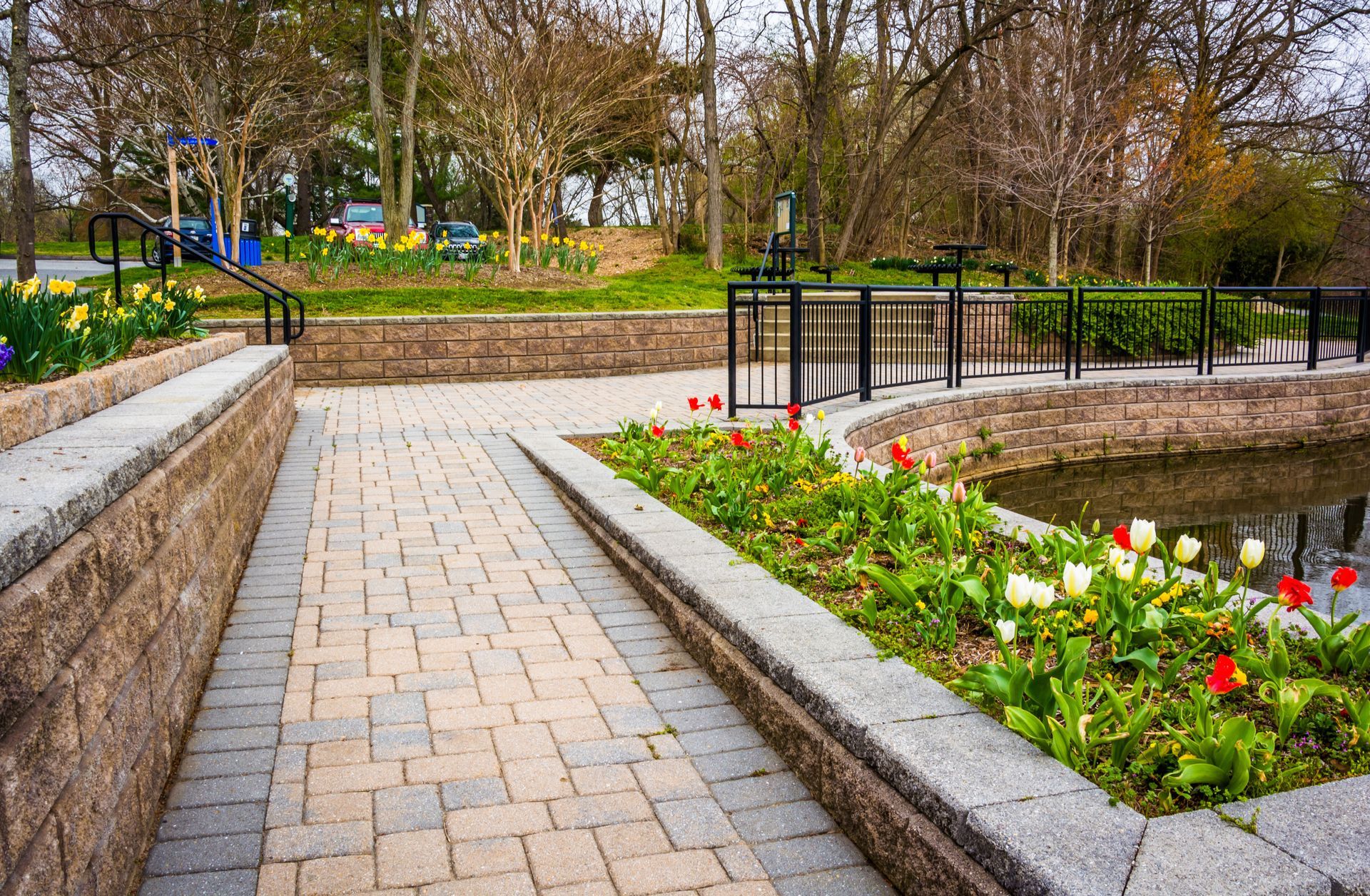 A paved stone walkway leads past a garden bed with tulips and a small pond, bordered by tiered stone retaining walls.