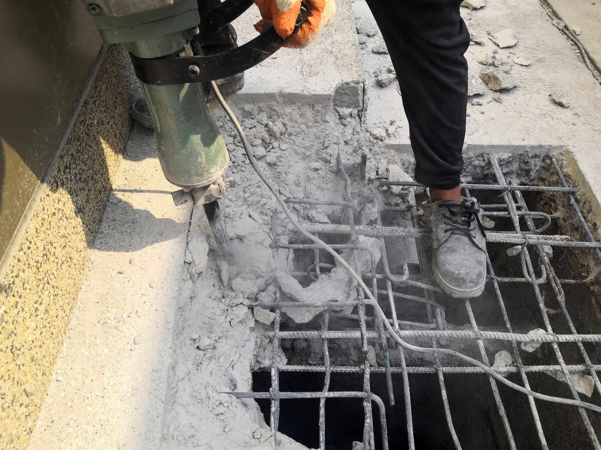 Person using a jackhammer to break concrete near a metal grate, creating dust.