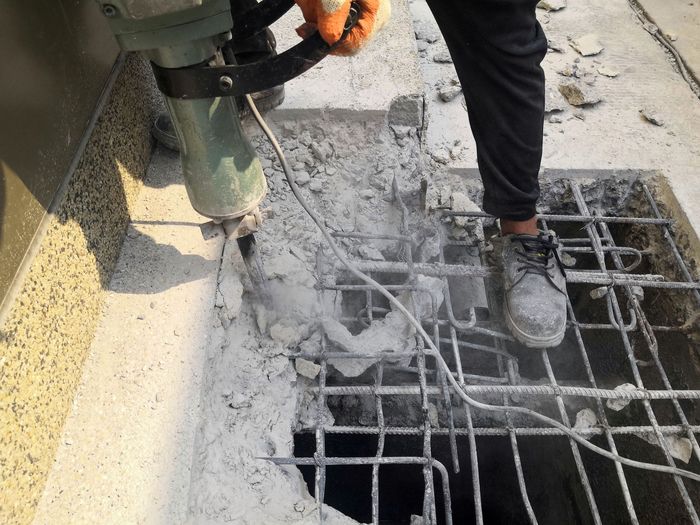 Person using a jackhammer to break concrete near a metal grate, creating dust.