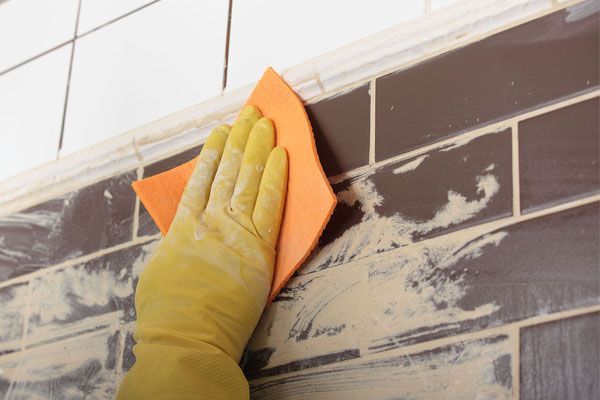 Gloved hand sanding brown wall tiles with orange sanding block near white upper tiles