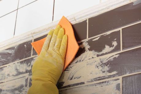 Gloved hand sanding brown wall tiles with orange sanding block near white upper tiles