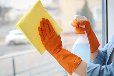 Learn More About Commercial Cleaning Person in orange gloves cleaning a window with a yellow cloth and spray bottle