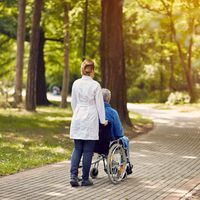 A woman pushes an elderly man's wheelchair through a tree-lined path.