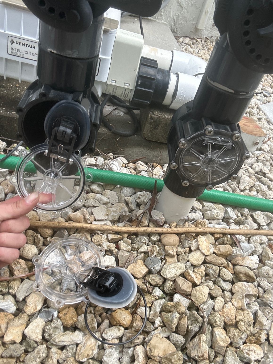 A person holds a clear, circular valve cover removed from a pool plumbing fixture near a second, installed valve cover.