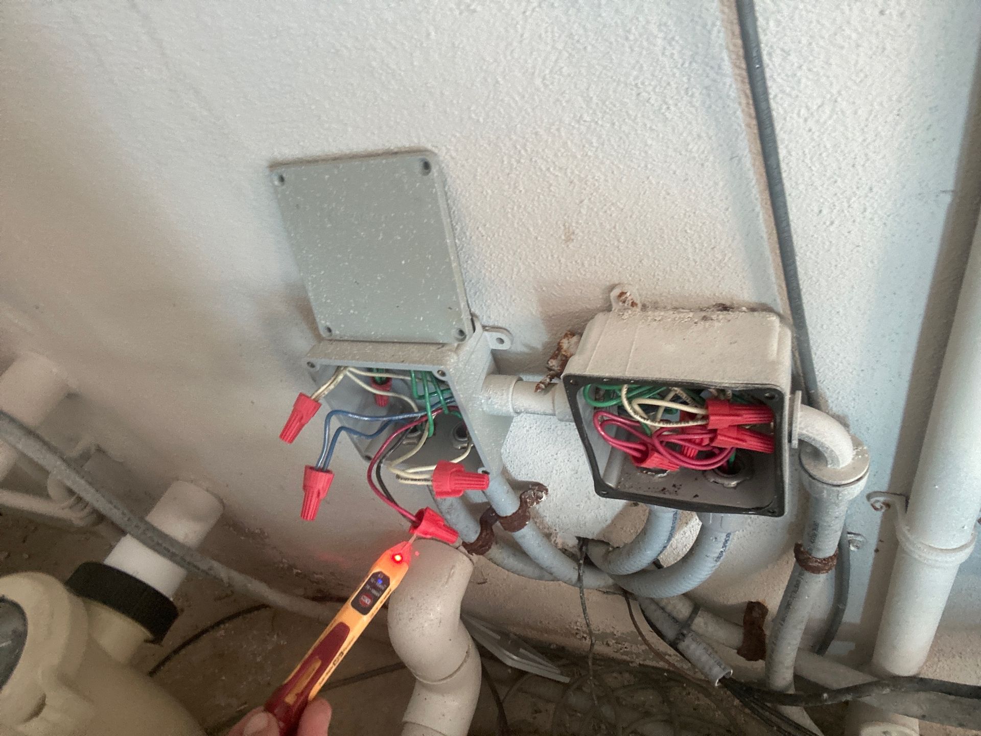 A hand holds a non-contact voltage tester against exposed electrical wires inside open junction boxes on a wall.