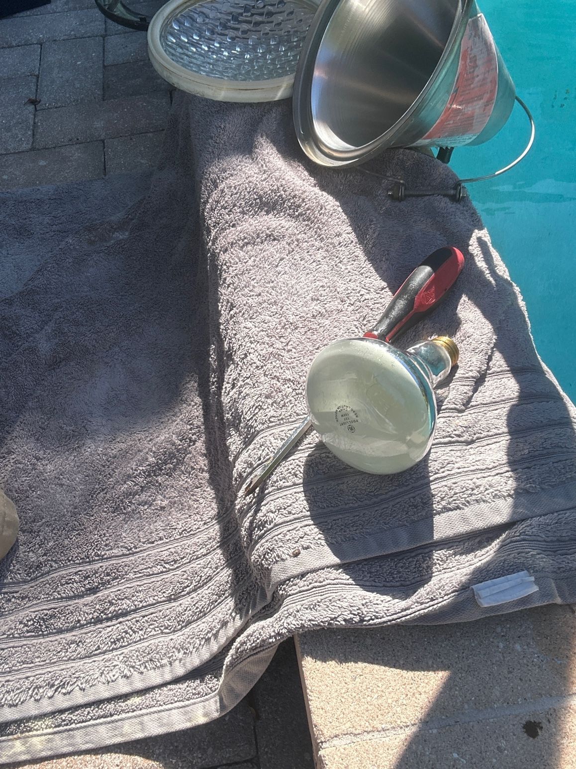 A disassembled swimming pool light and a screwdriver resting on a towel next to a pool.