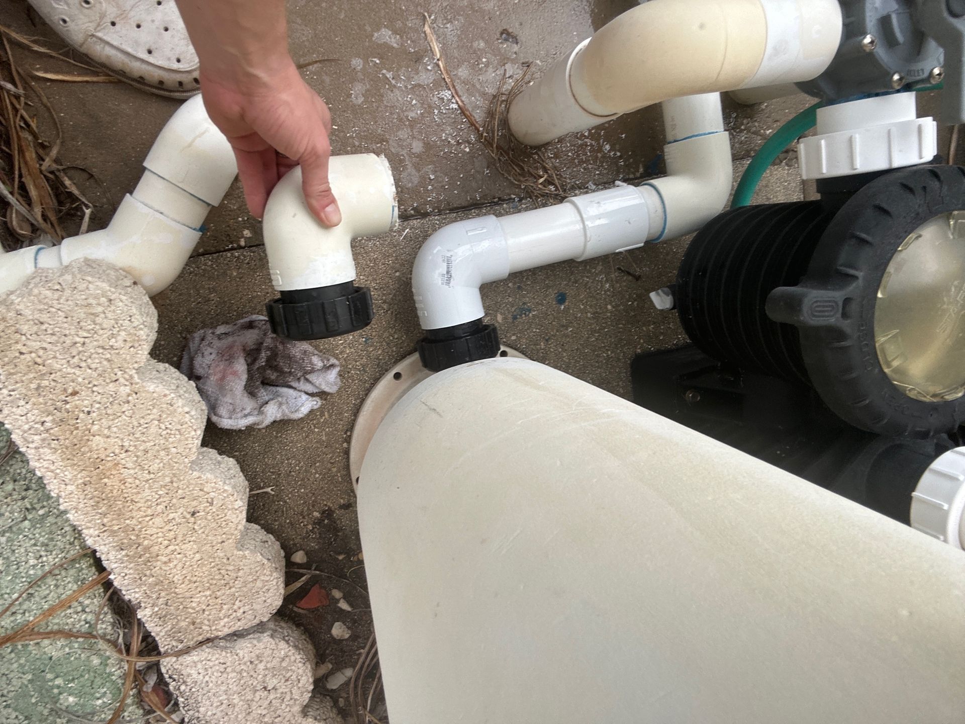 A hand holds a disconnected PVC pipe elbow near the plumbing fixtures of an outdoor pool filter system.