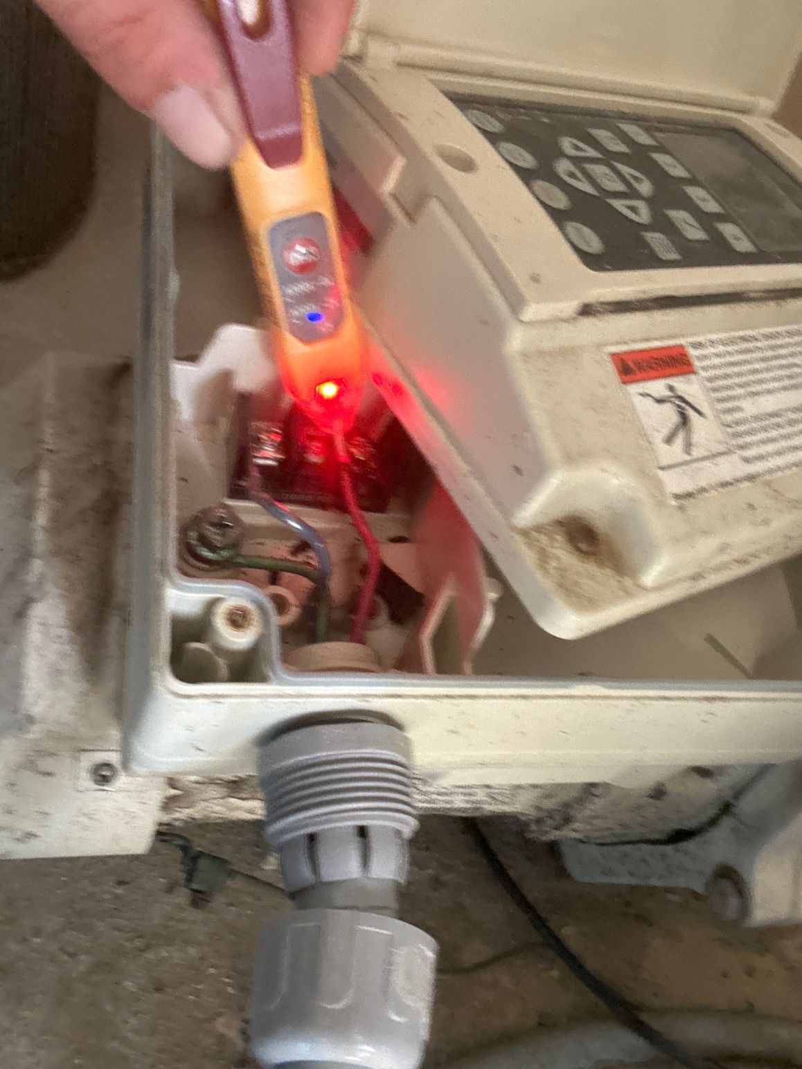 A hand uses a non-contact voltage tester to check a red wire inside an open electrical control box.