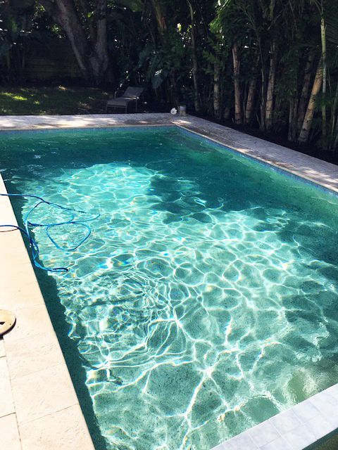 A rectangular swimming pool filled with clear, turquoise water, surrounded by light-colored stone tiling and lush trees.