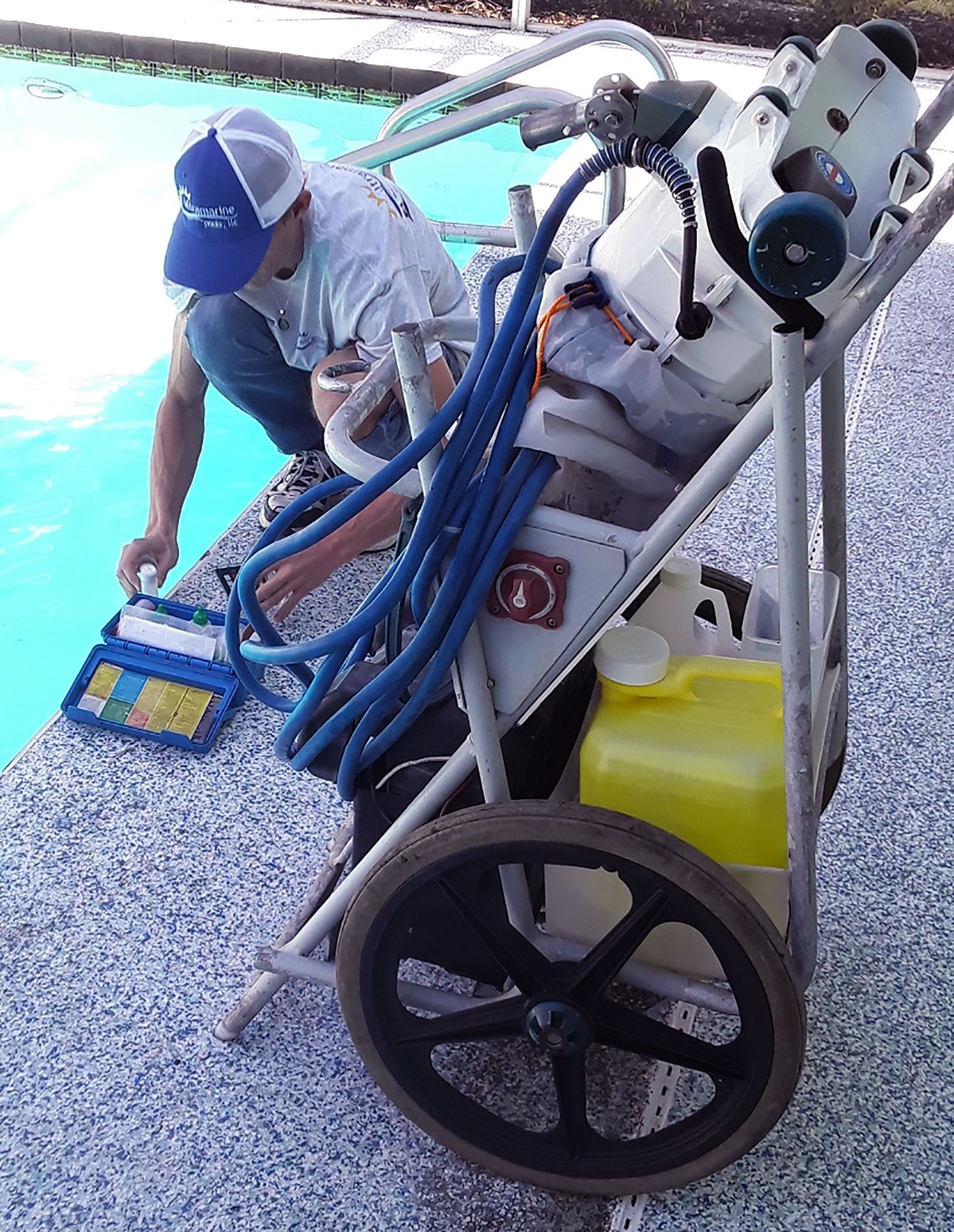 A technician in a blue hat tests pool water chemistry using a test kit next to a portable pool service cart.
