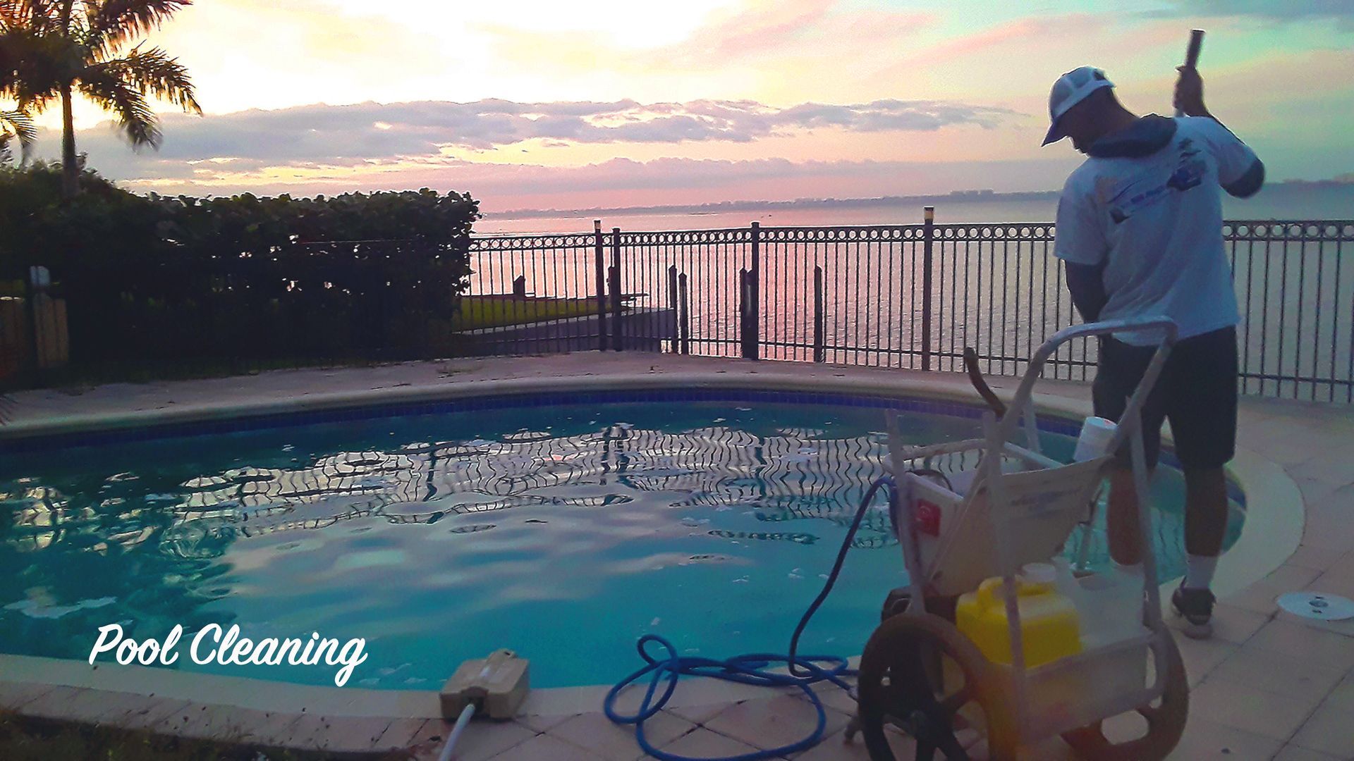 A technician uses cleaning equipment by a backyard swimming pool at sunset, with a fence and water in the background.