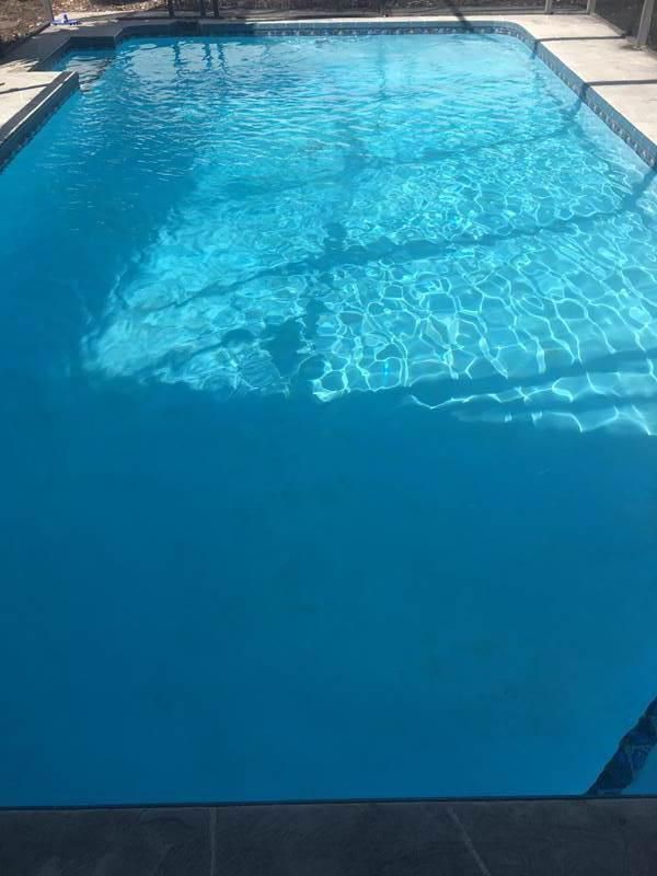 A bright blue backyard swimming pool surrounded by a concrete deck with dark shadows cast across the water.