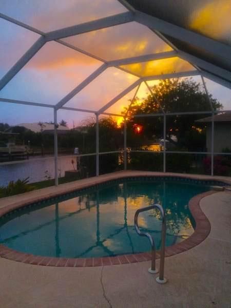 A sunset viewed through a screened pool enclosure, with vibrant pink and gold clouds reflecting on the water.