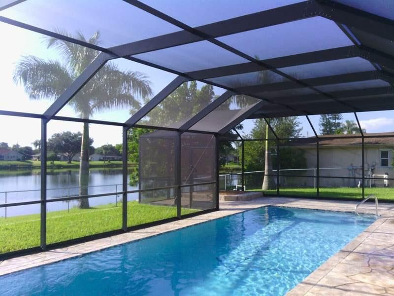 A swimming pool enclosed by a dark aluminum screen cage, with a view of a grassy lawn, a lake, and palm trees beyond.