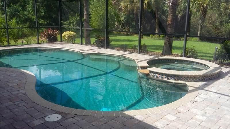A blue swimming pool with an attached circular hot tub, enclosed by a screen patio overlooking a grassy backyard.