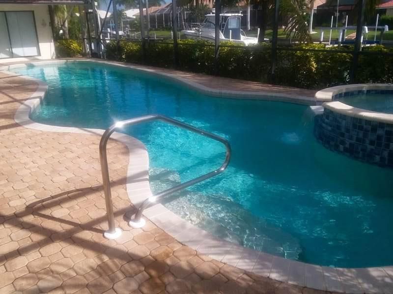A swimming pool with curved edges, a metal handrail at the steps, and an adjacent spa, set on a tan brick patio.