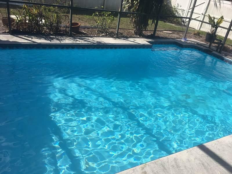 A bright blue swimming pool framed by a patio and a screened-in enclosure with potted plants in the background.