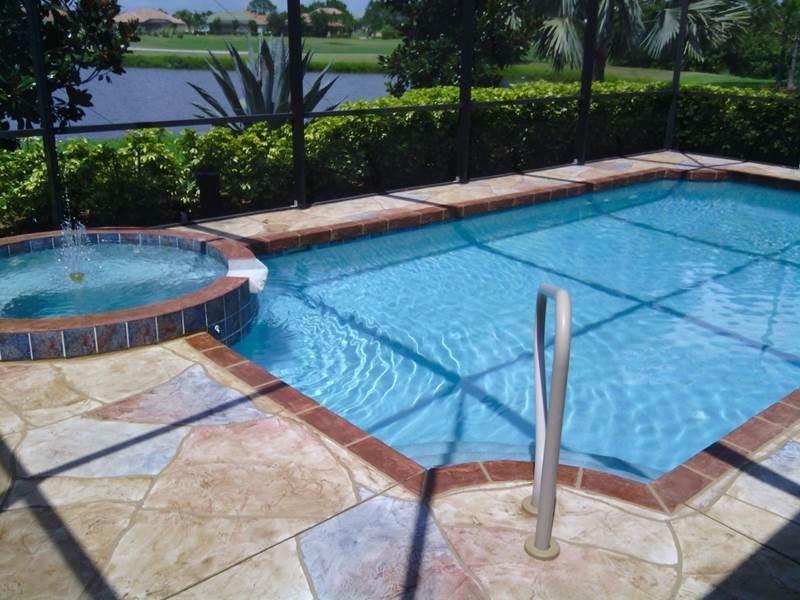 A blue swimming pool and attached circular spa on a stone deck, enclosed by a screen, overlooking a lake and greenery.