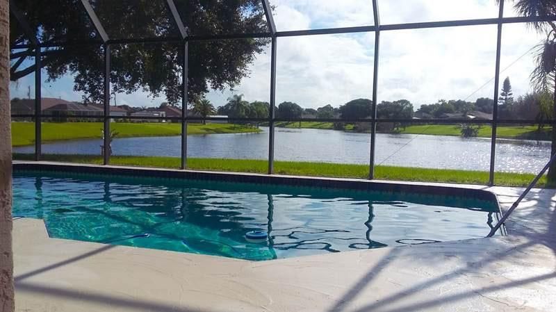 A swimming pool sits on a patio enclosed by a screen, overlooking a lake surrounded by green grass and houses.