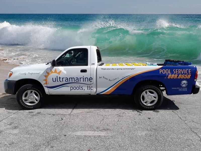 A white and blue Ultramarine Pools, LLC service truck parked on a beach in front of large crashing waves.