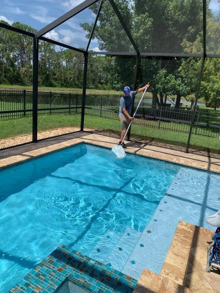 Person skimming a blue swimming pool inside a screened enclosure, with trees and a fence beyond.
