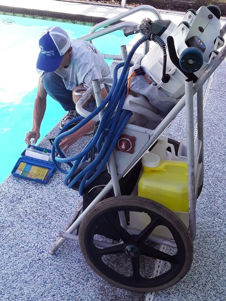 Worker on a pool-cleaning cart vacuuming a pool edge with hoses and equipment attached