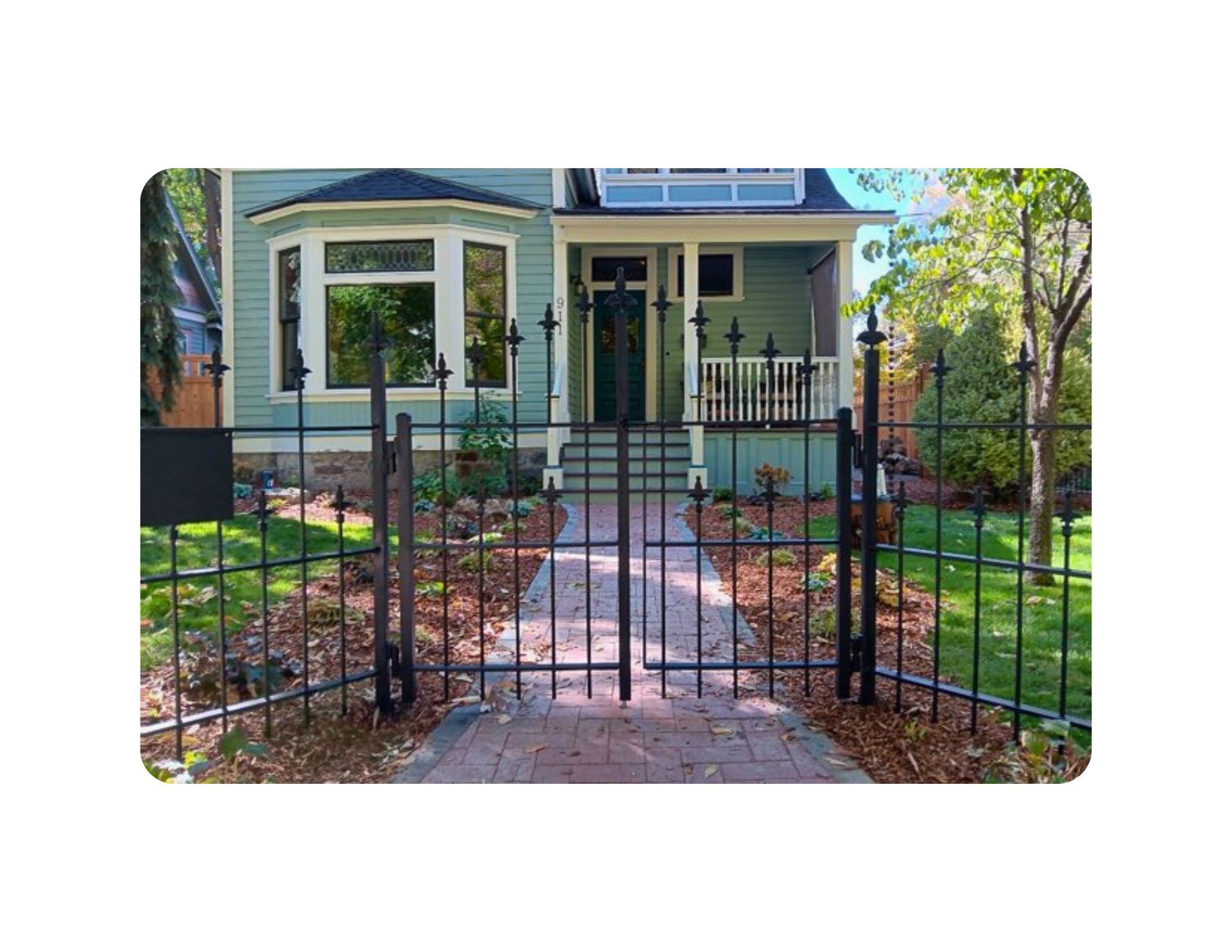 Black wrought iron gate leading to a light blue house with a porch and windows, a brick pathway, and a grassy yard.