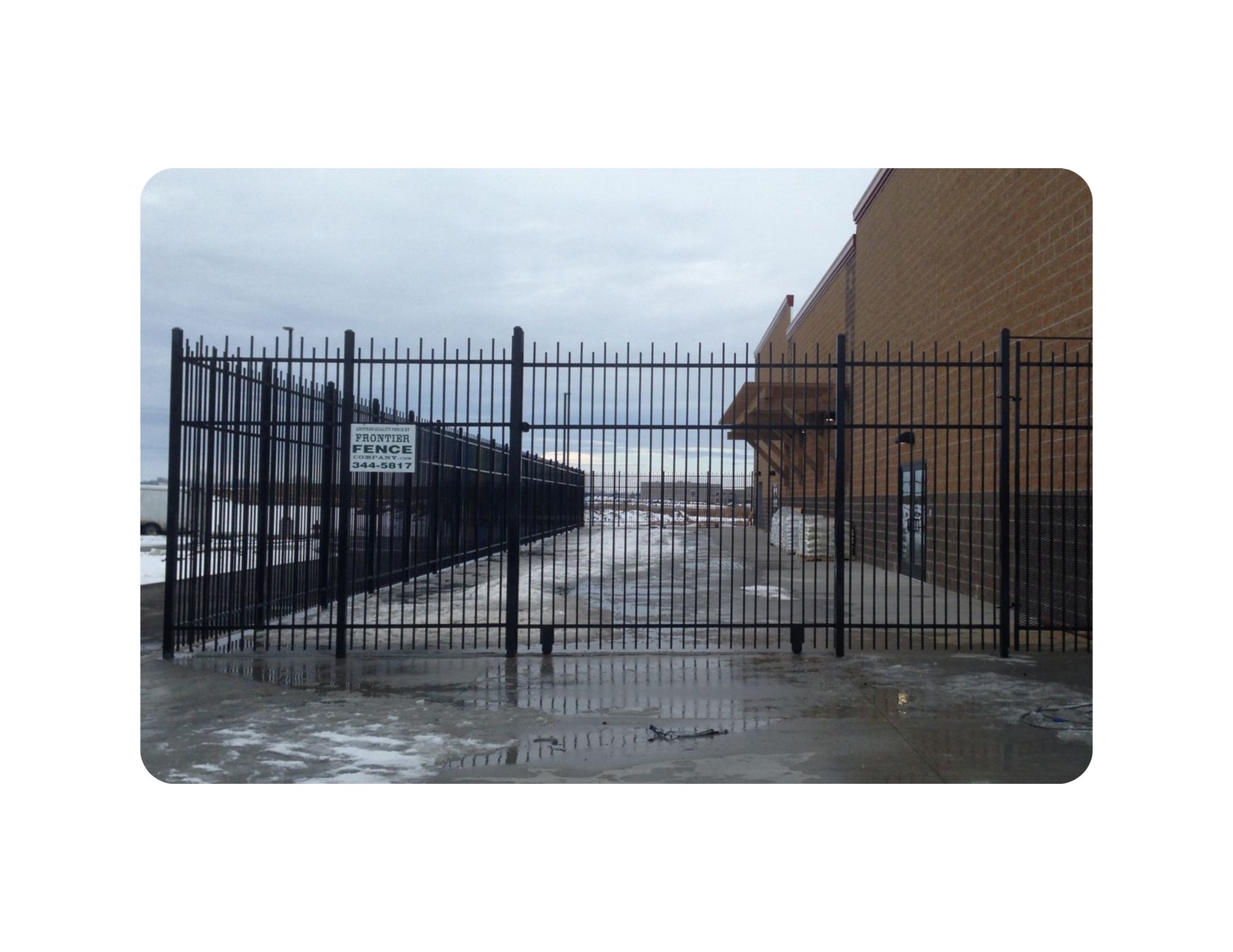 Black metal fence with gate, near building, water visible, cloudy sky.