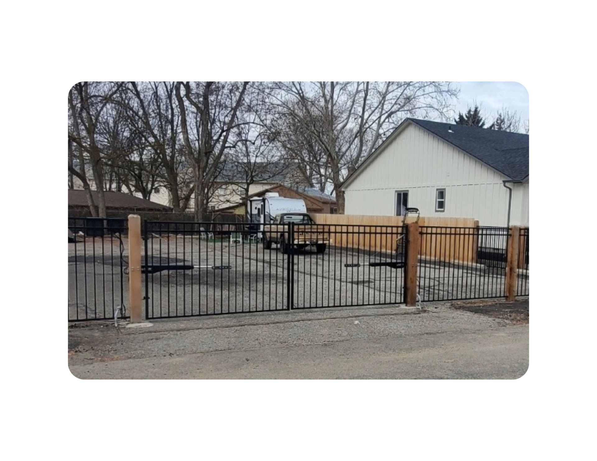 Black metal gate with wooden posts; gravel driveway, beige building in the background.