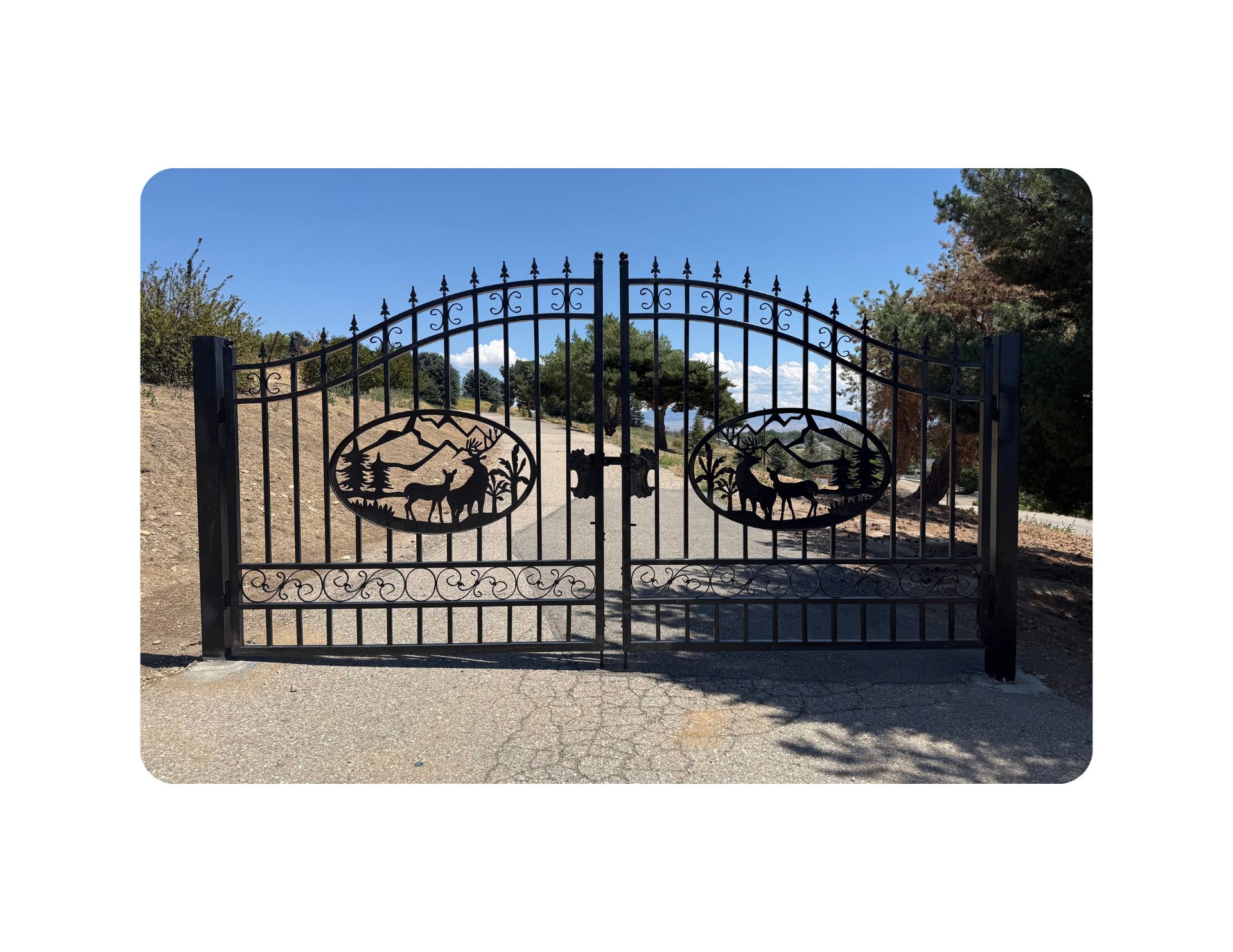 Black wrought iron gate with deer silhouettes, set in a gravel drive under a blue sky.
