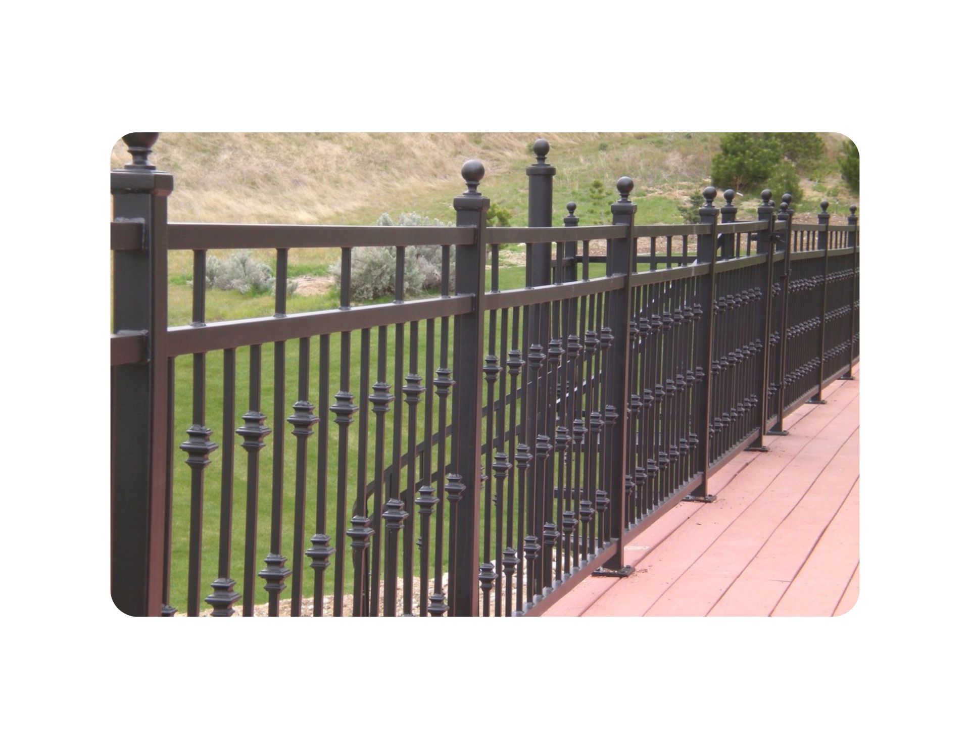 Black metal fence on a wood deck, with decorative vertical bars. Background includes grass and a hillside.
