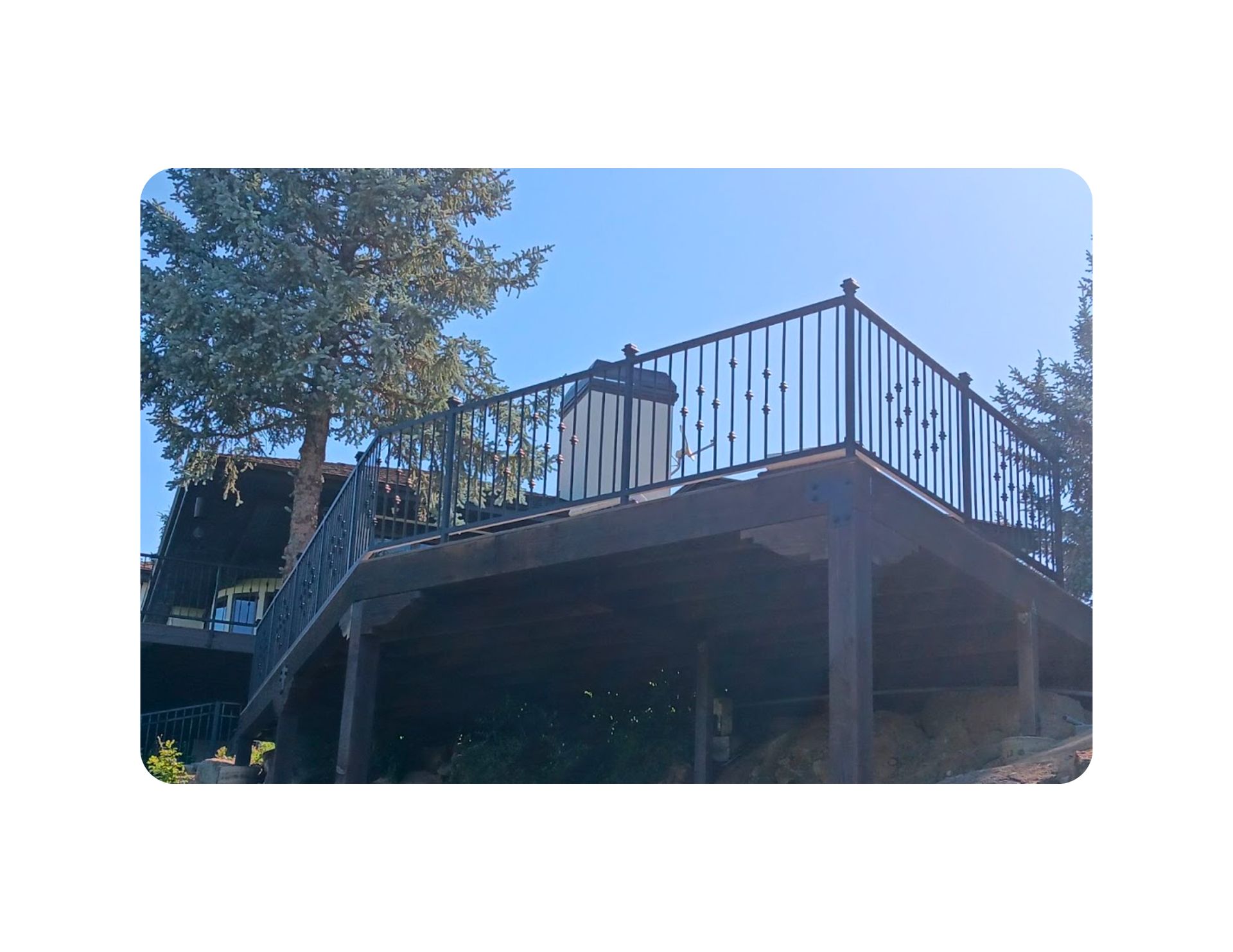 Dark brown deck with black railing against a bright blue sky, tall tree.
