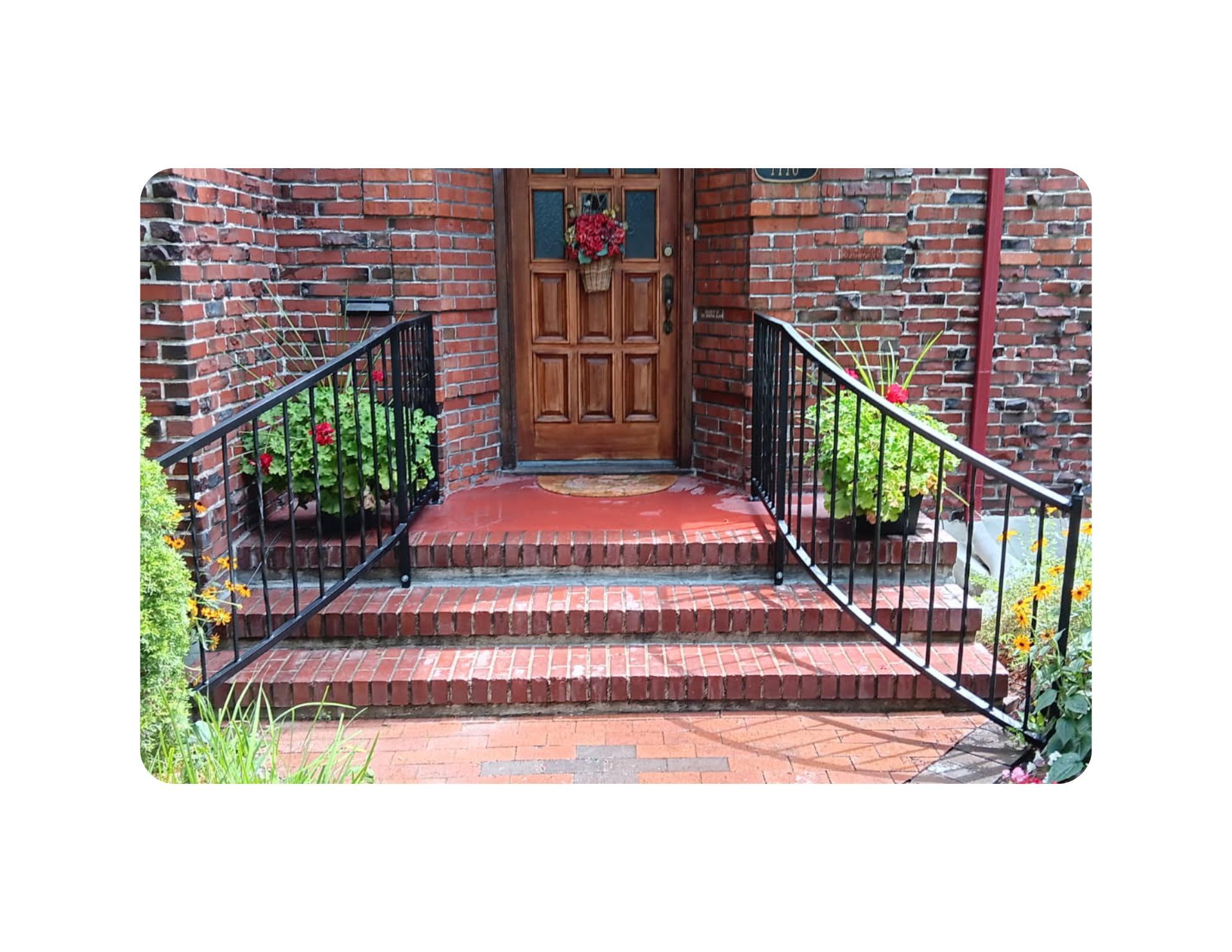 Brick steps leading to a wooden door with a floral wreath; black railings and potted plants flank the entrance.