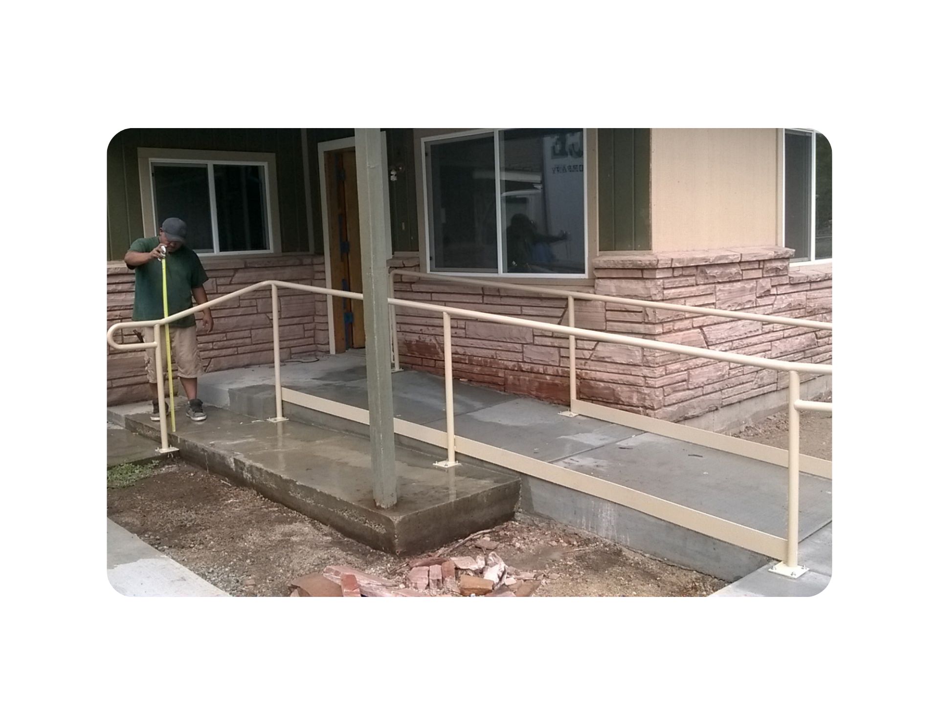 Ramp with handrails leading to a building entrance. A person waters the ground with a hose.