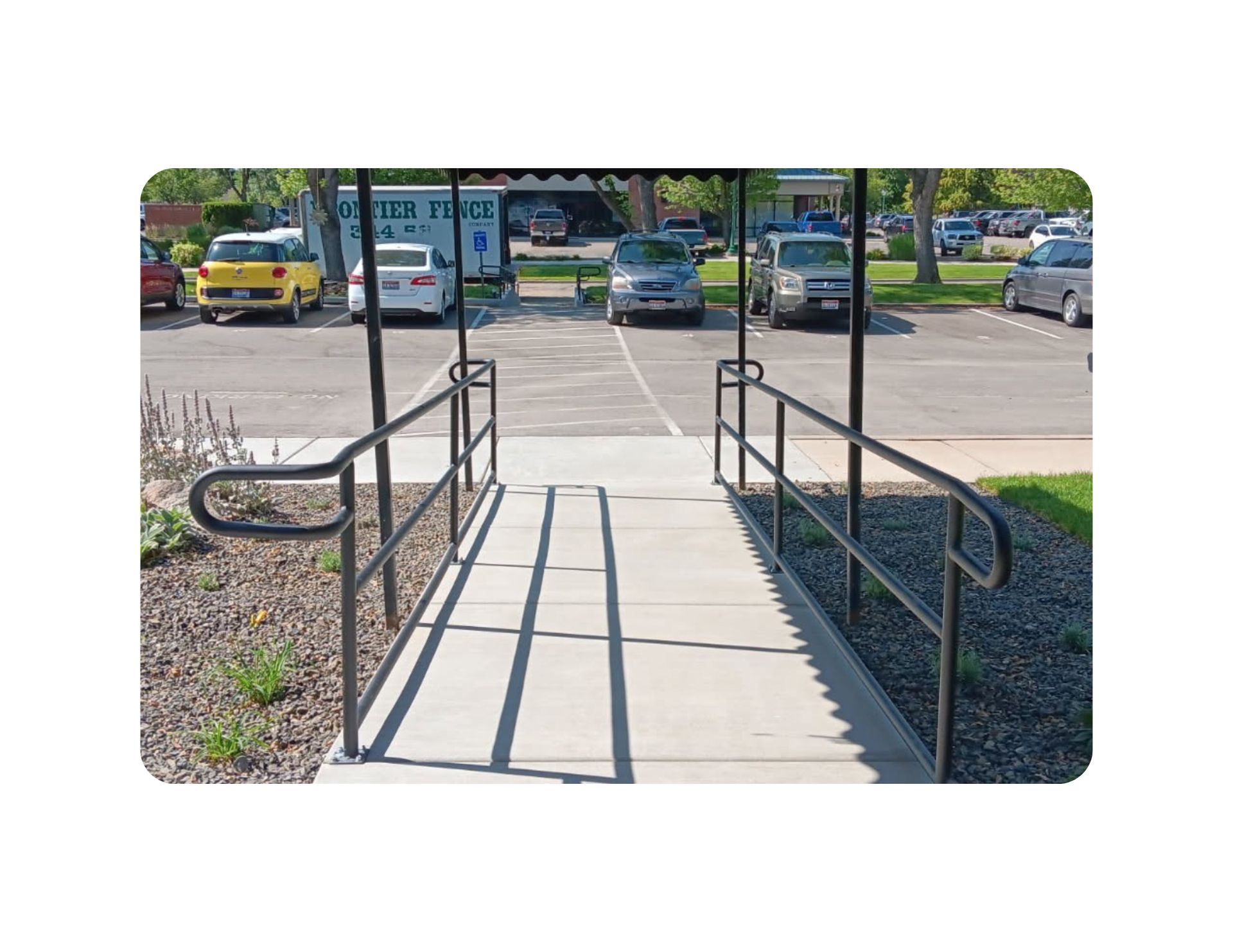 Ramp with black handrails, leading to a covered entrance. Cars parked in the background.