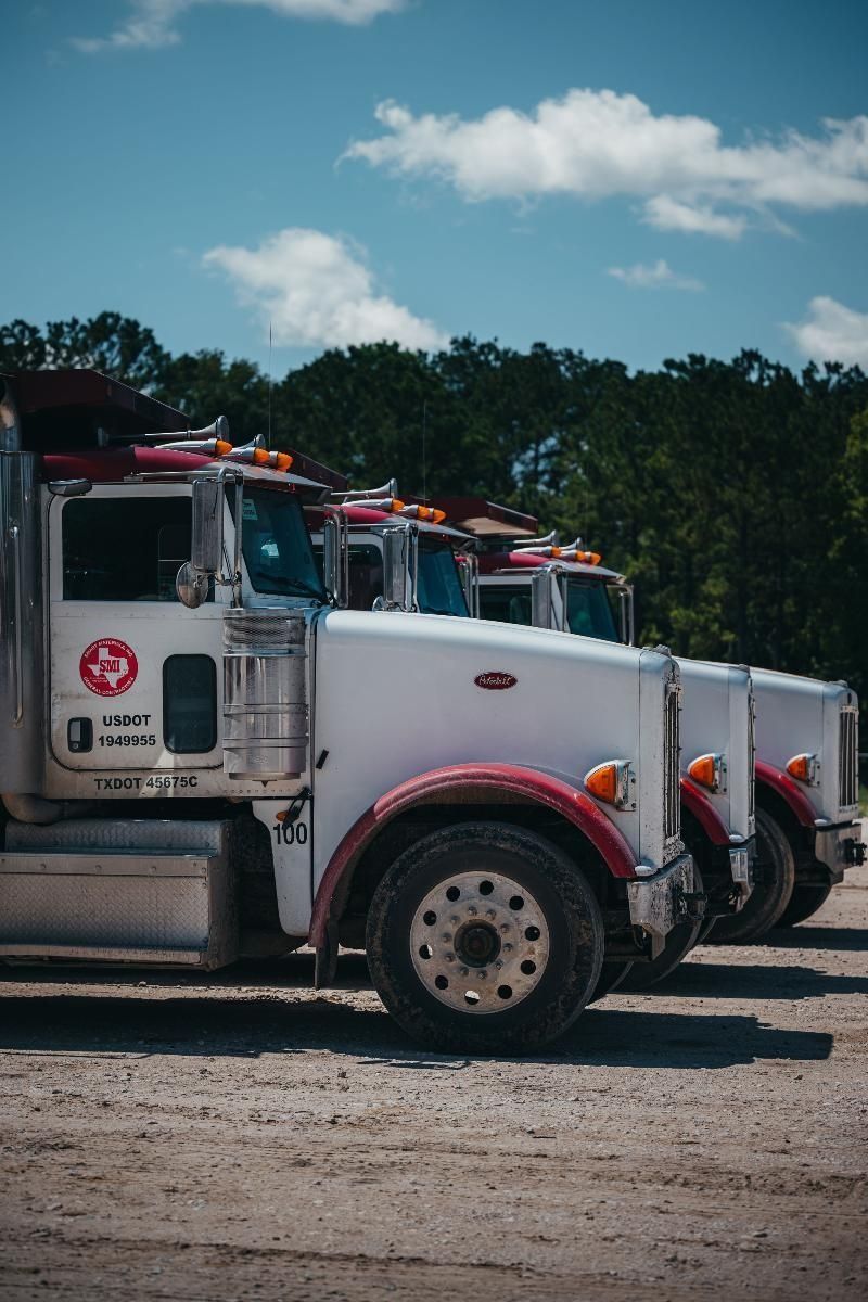 Three white Peterbilt semi-trucks parked in a row on a dirt lot, against a blue sky with trees in the background.