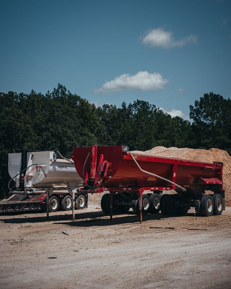 Two semi-trailers on a dirt lot; one red filled with wood chips, one silver. Trees and blue sky background.