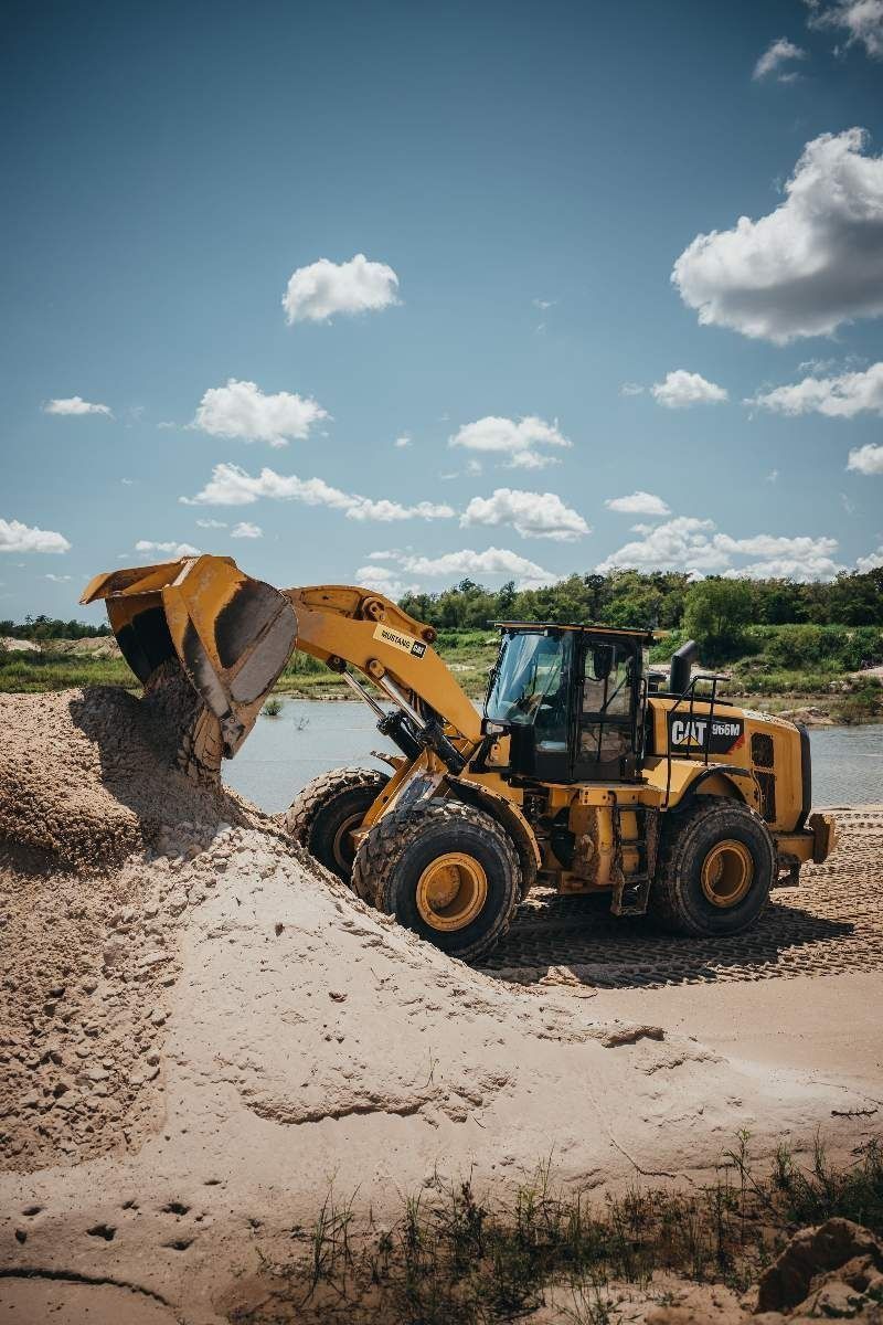 Yellow CAT wheel loader scoops sand by a river under a blue sky.
