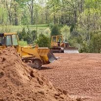 Bulldozers on dirt clearing land, with a pile of dirt in the foreground and trees in the background.