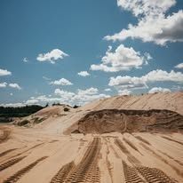 Sandy dunes under a blue sky with scattered clouds. Tire tracks cross the foreground.