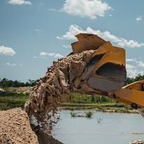 Excavator bucket dumping dirt into a body of water. Sunny day with blue sky and white clouds.