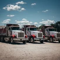 Three red and white dump trucks parked on a dirt lot under a cloudy blue sky.