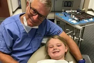 A dentist is sitting next to a little girl in a dental chair.