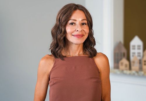 Woman standing indoors, smiling in a rust-colored sleeveless top with a blurred shelf in the background