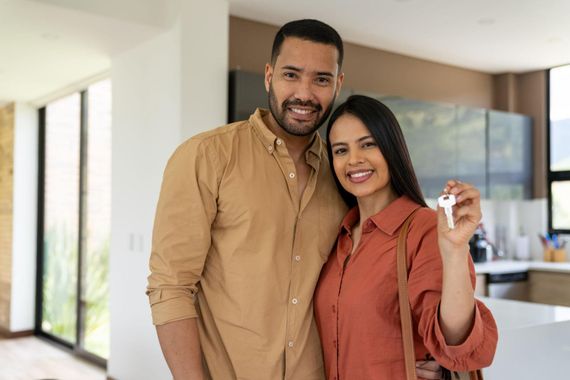 Smiling couple standing indoors, holding up house keys in a bright living room