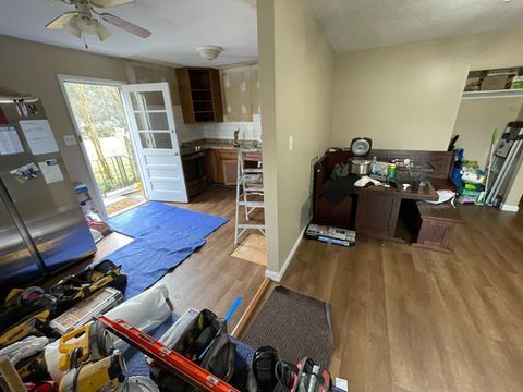 Cluttered kitchen and living area with open door, blue rug, and scattered tools and furniture